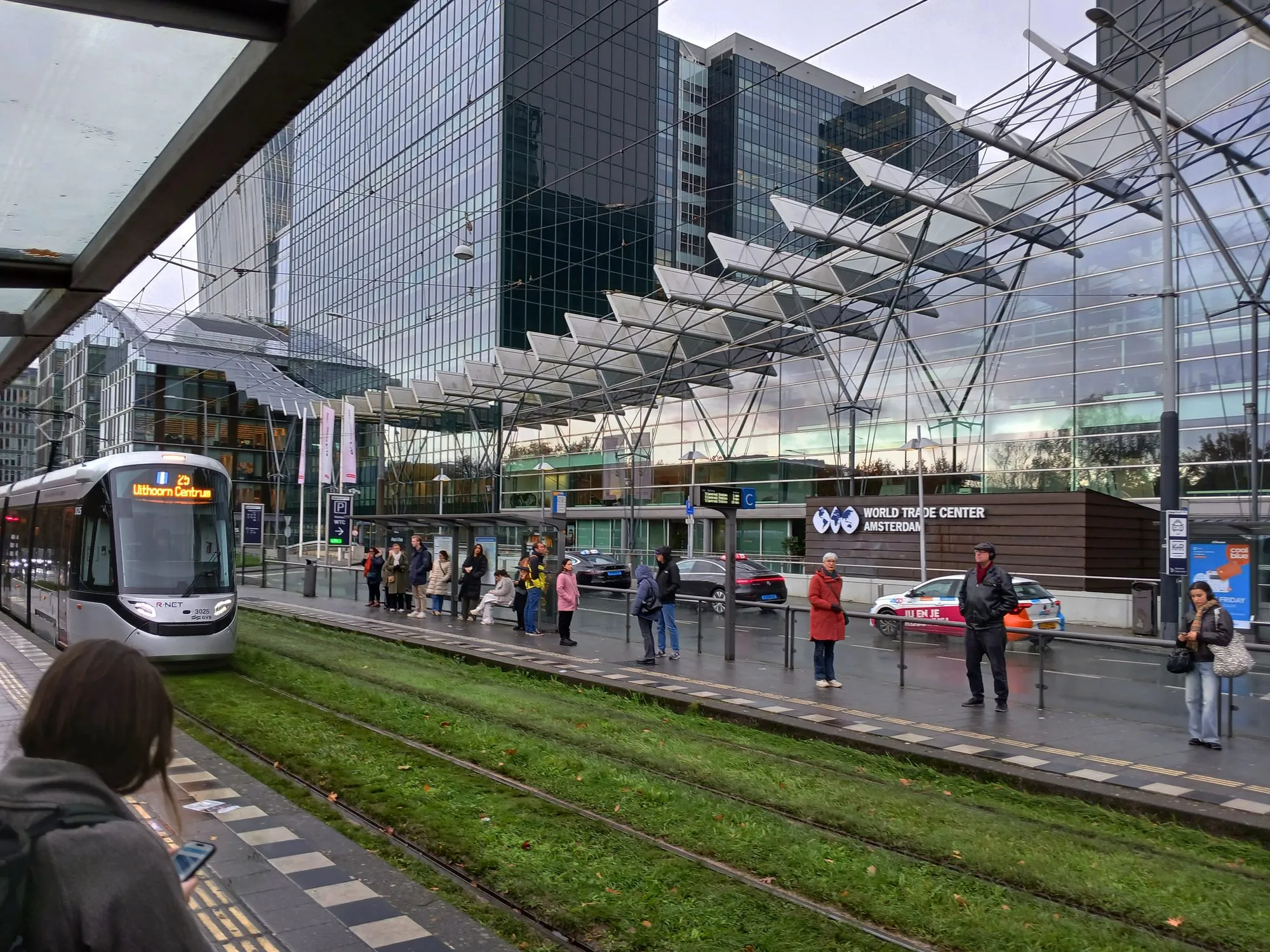 Oameni care așteaptă la o stație de tramvai lângă gara Amsterdam Zuid, cu turnurile de birouri din sticlă ale districtului de afaceri Zuidas și World Trade Center Amsterdam în spate