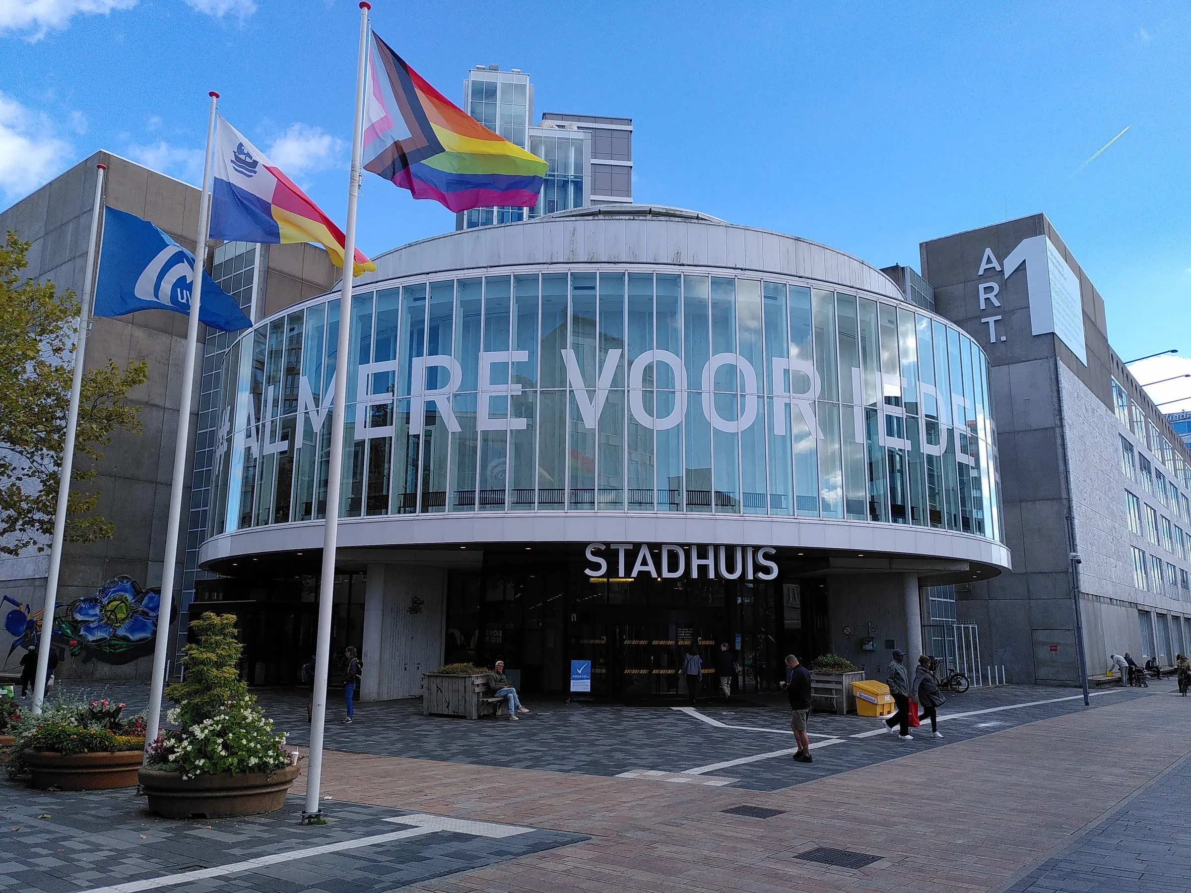 The rounded glass front of Almere city hall on Stadhuisplein, with the words 'Almere voor iedereen' across the windows and flags flying in front