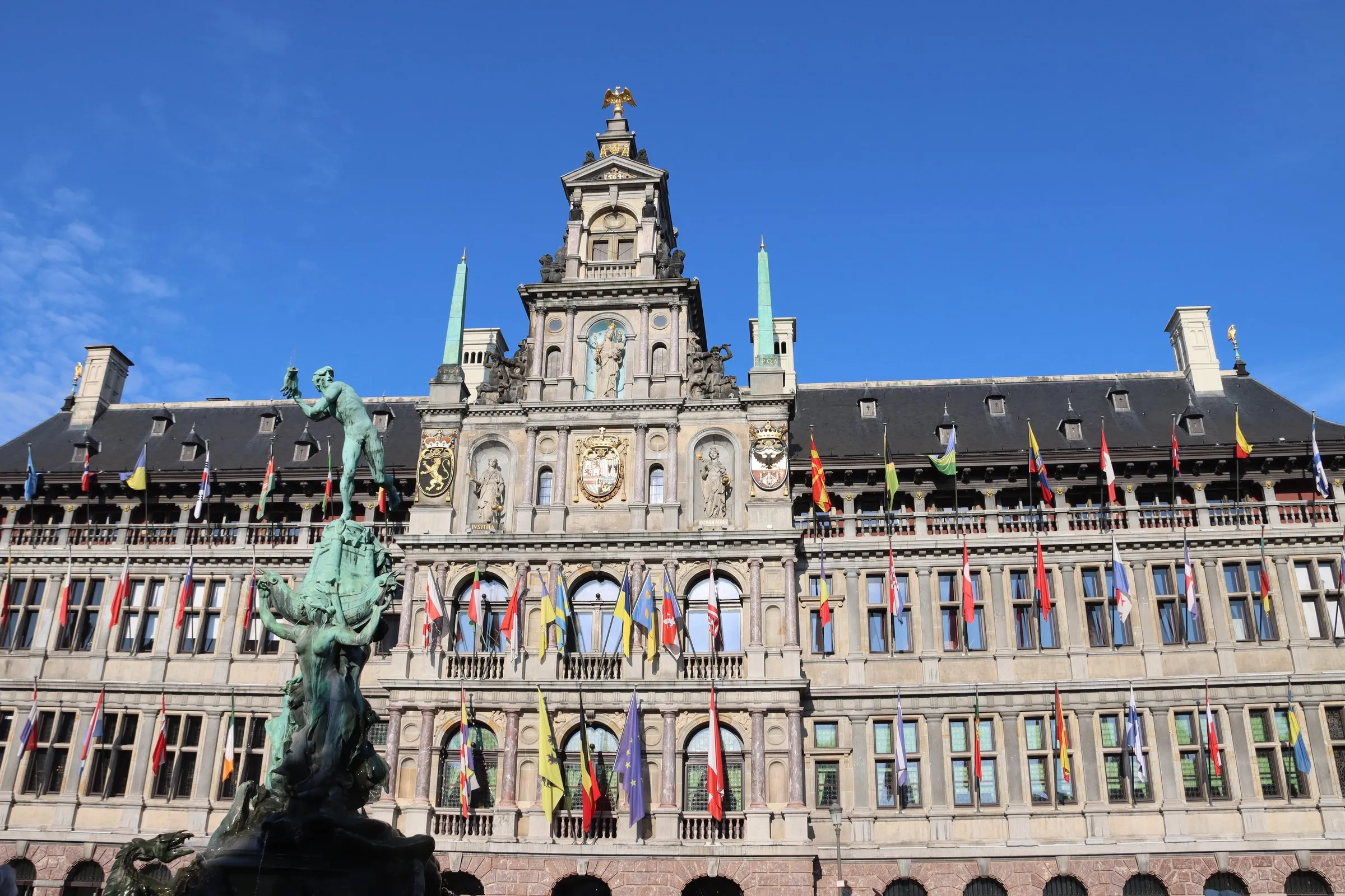 Antwerp City Hall on the Grote Markt, its Renaissance facade hung with rows of national flags, with the bronze Brabo fountain in front under a blue sky