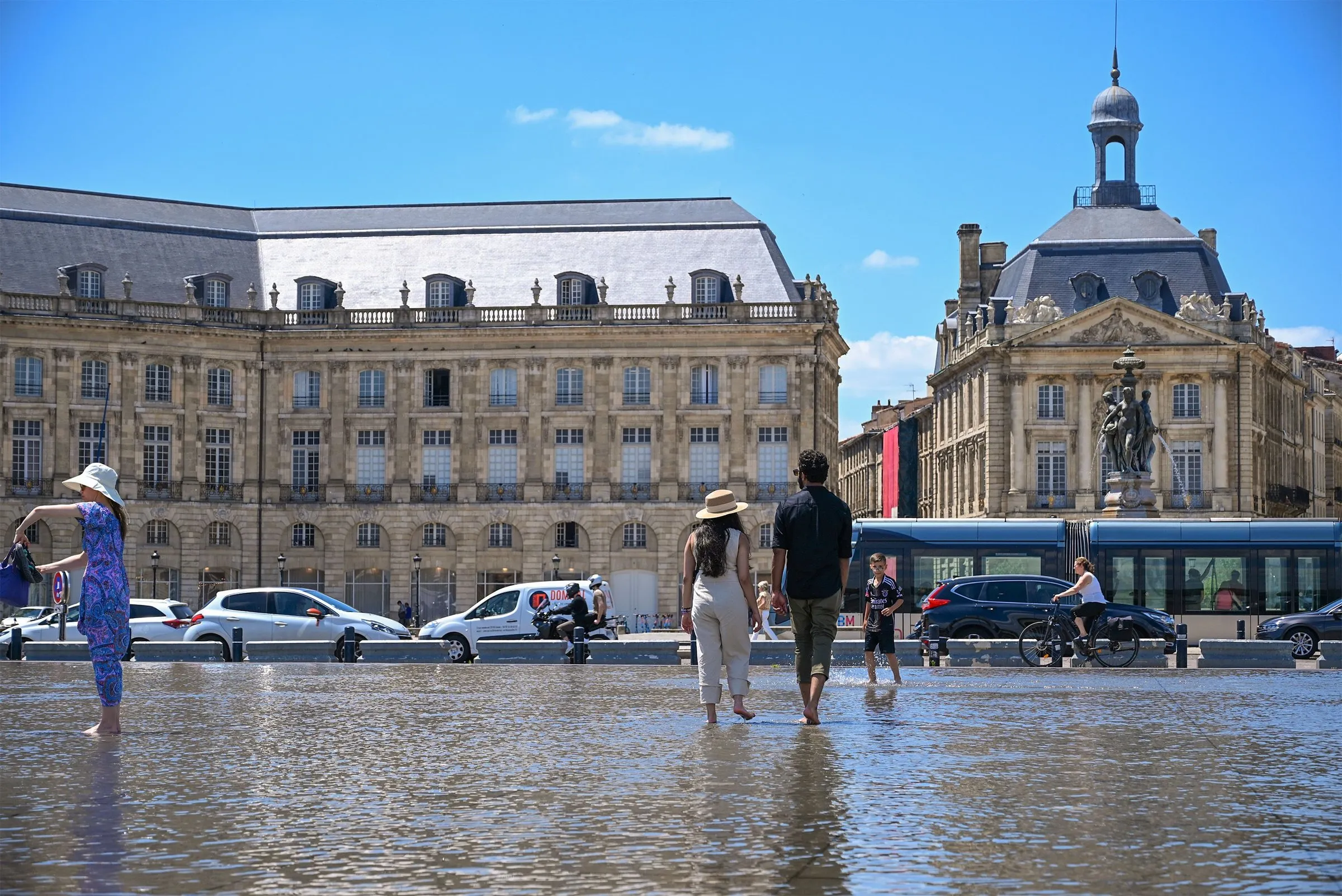 People wading across the Miroir d'eau on Place de la Bourse in Bordeaux on a sunny day, with a tram, cars and a cyclist passing in front of the stone façades and the Three Graces fountain