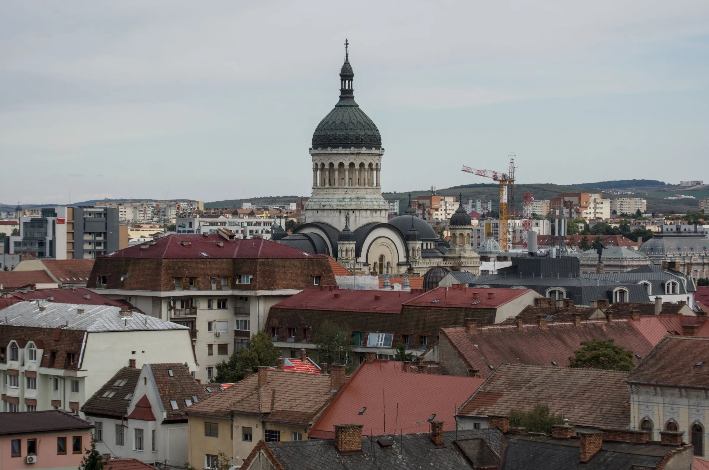 Rooftops of central Cluj-Napoca with the dome of the Orthodox Metropolitan Cathedral, newer buildings, a construction crane and hills behind