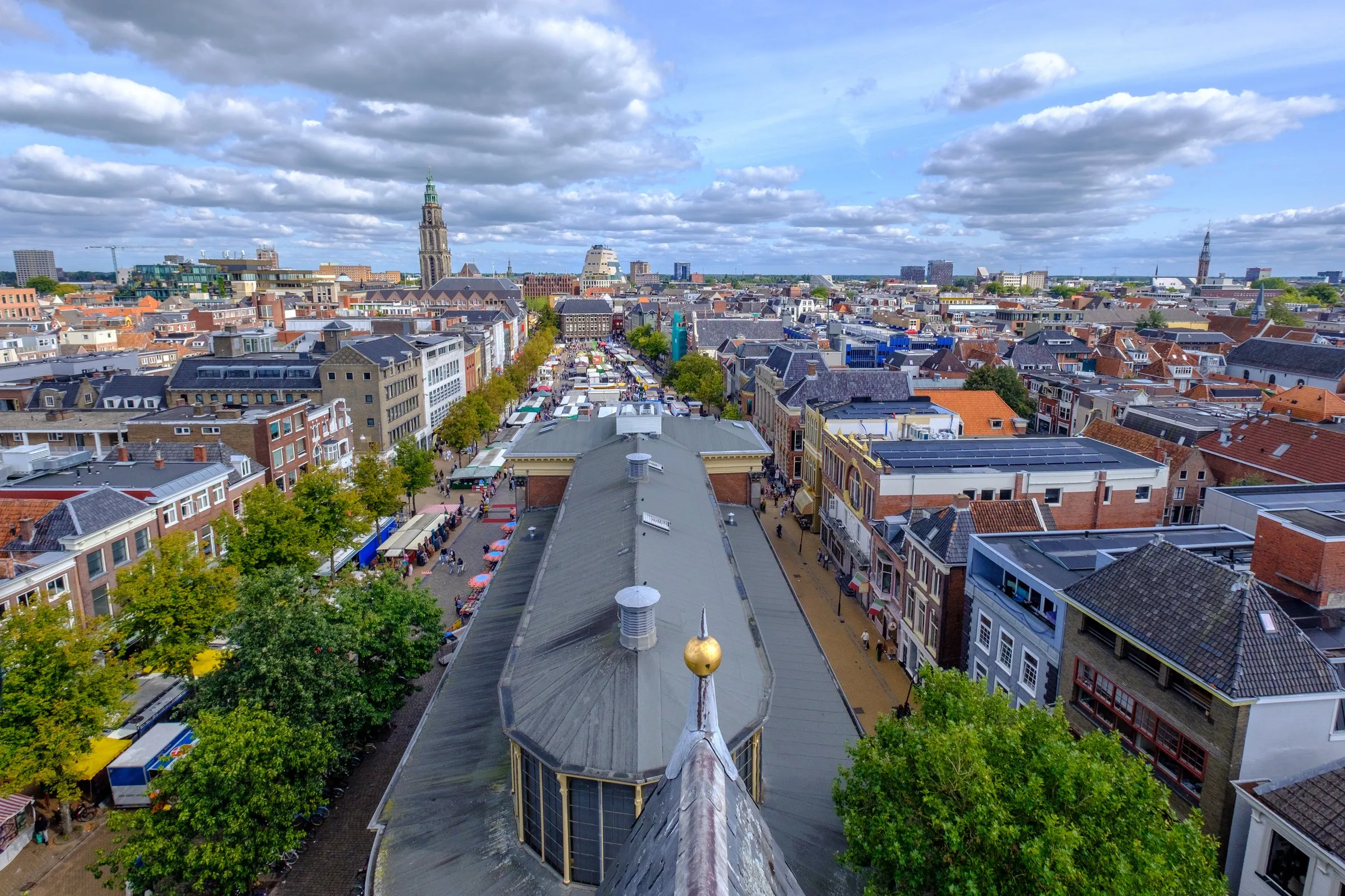 View from the vaults of the Der Aa-kerk over the grey roof of the Korenbeurs and the market stalls of the Vismarkt, with the Martinitoren rising above the rooftops of central Groningen under a cloudy sky
