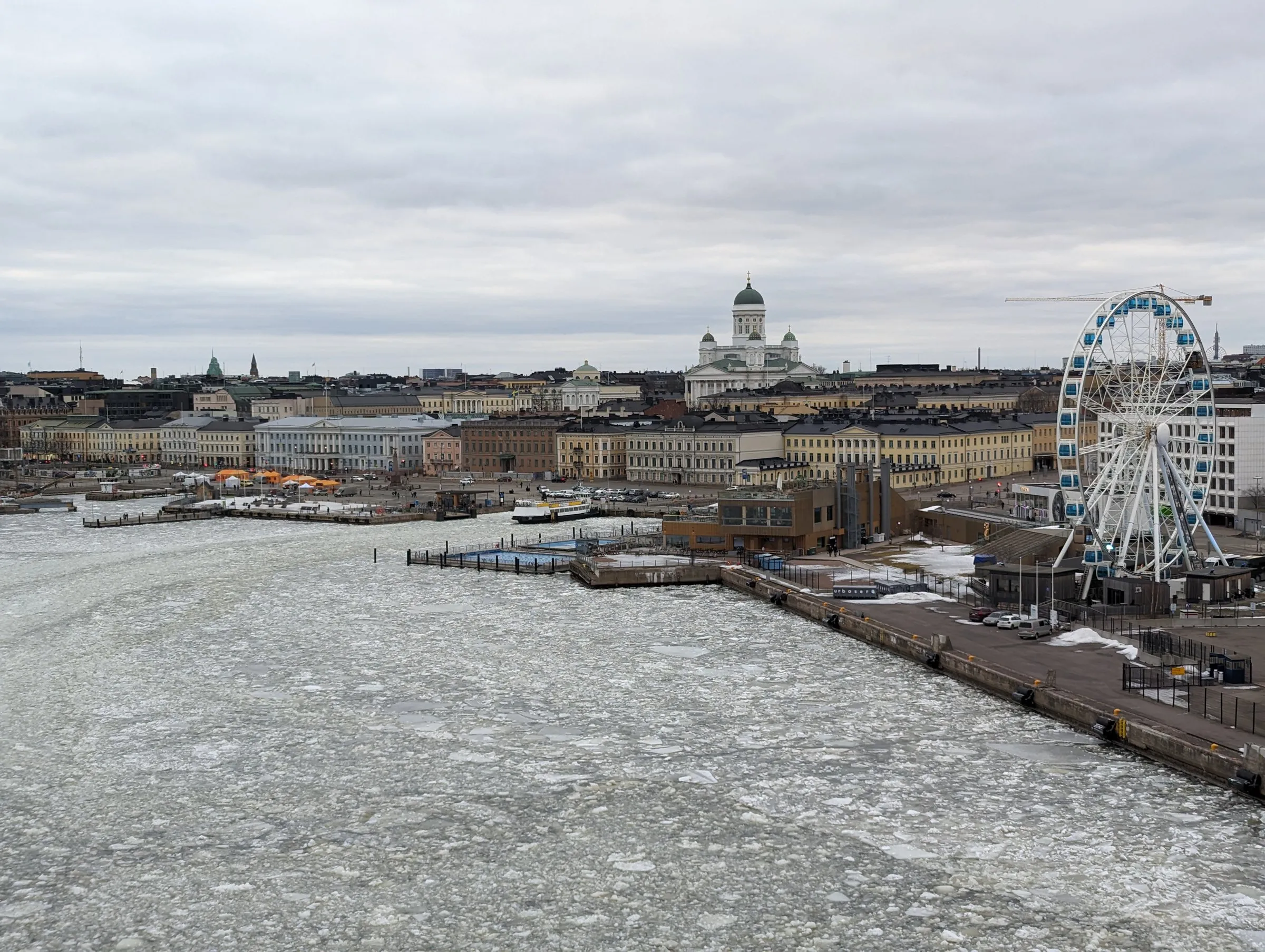 Helsinki's South Harbour covered in broken ice under a grey sky, with the white Helsinki Cathedral rising above the waterfront buildings and the SkyWheel Ferris wheel on the right