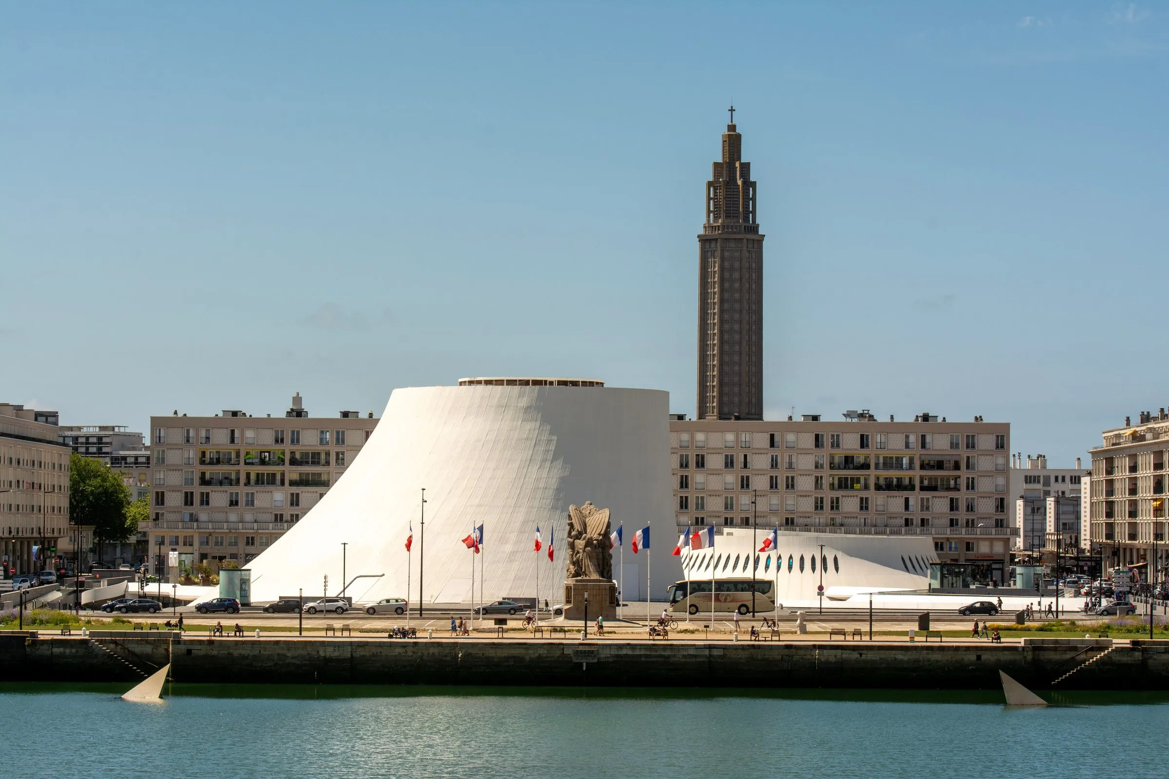 Le Havre's waterfront on a sunny day: the white, curving Volcan cultural centre by Oscar Niemeyer and a war memorial with French flags in front of post-war apartment blocks, with the concrete tower of St Joseph's Church rising behind and a harbour basin in the foreground