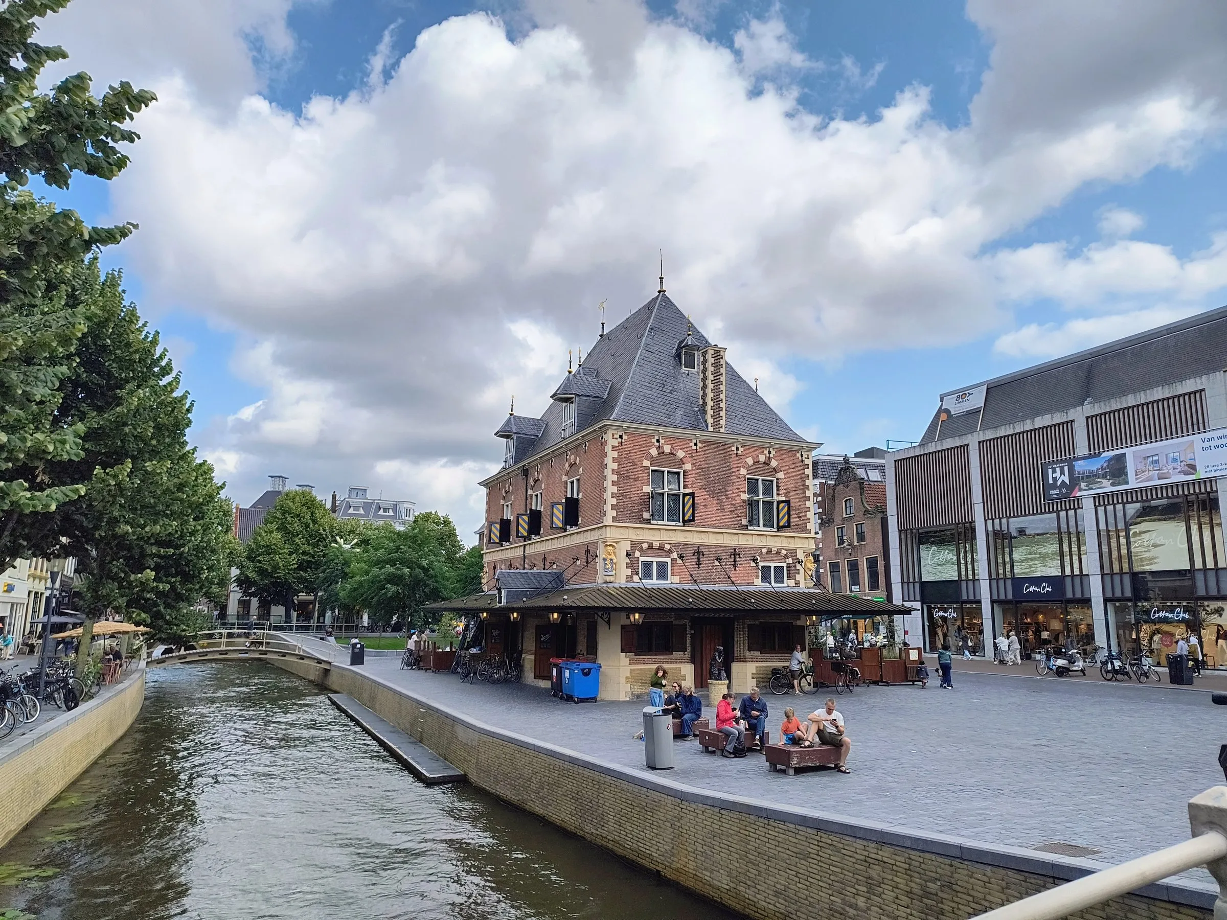 The Waag, a red-brick weigh house with a steep slate roof and striped shutters, beside a canal in central Leeuwarden, with people sitting on benches on the square and a modern shopping building on the right