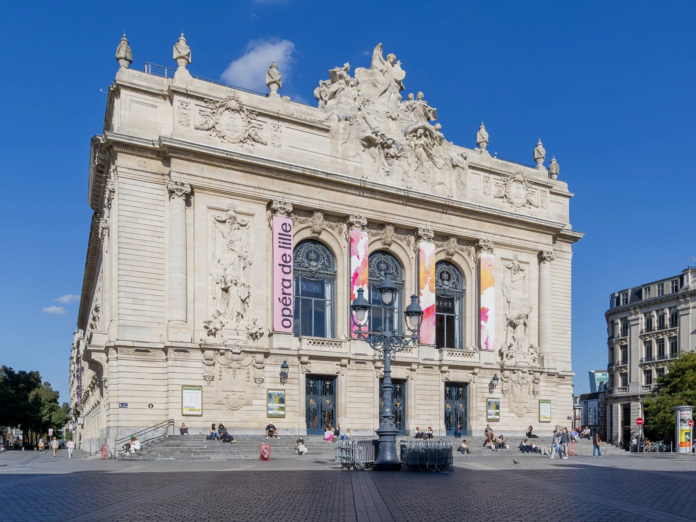 The Opéra de Lille under a deep blue sky: a pale stone neo-classical facade crowned by a sculpted pediment group, three tall arched windows hung with an "opéra de lille" banner and colourful posters, an ornate cast-iron lamp post in front, and people sitting on the broad front steps of the paved square