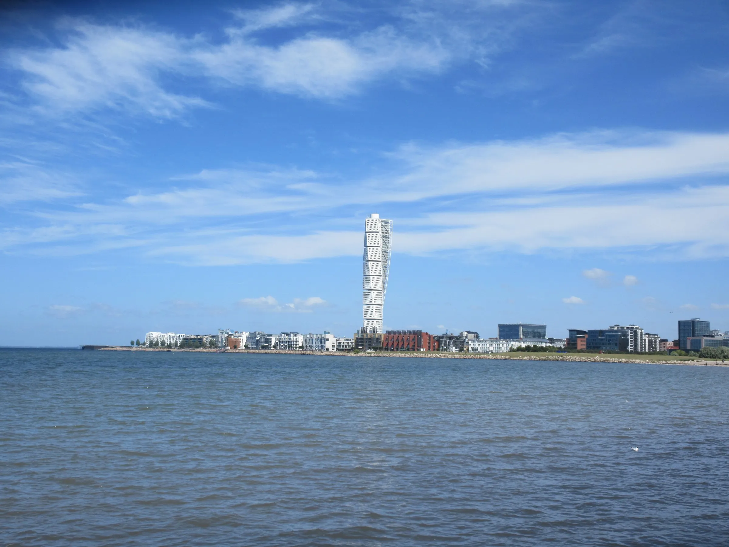 Turning Torso rising above the low waterfront blocks of Västra Hamnen, seen across the sea from Ribersborg beach under a blue sky with light clouds