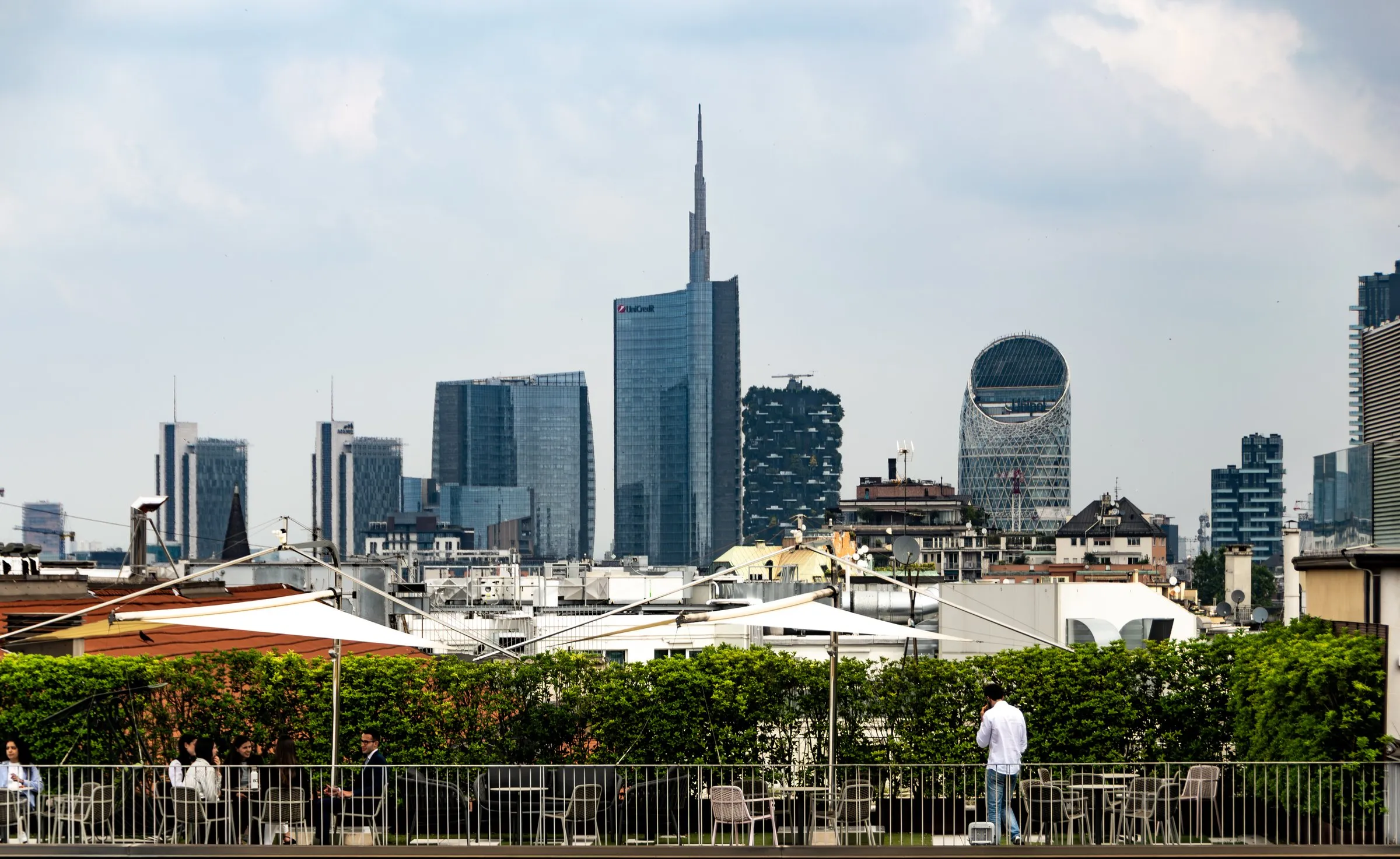 The Porta Nuova skyline in Milan under a cloudy sky, with the spire of the UniCredit Tower, the plant-covered Bosco Verticale towers and an oval lattice tower, seen from a rooftop terrace where people sit at tables