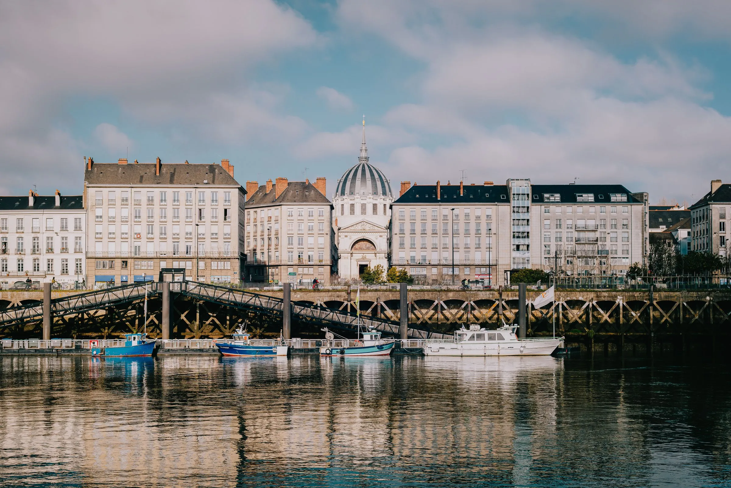The Loire in Nantes on a bright day with scattered clouds: a row of pale stone quayside buildings with grey slate roofs and a white church dome topped by a spire, a metal footbridge leading down to a pontoon where small fishing boats and a white motorboat are moored, all reflected in the calm water