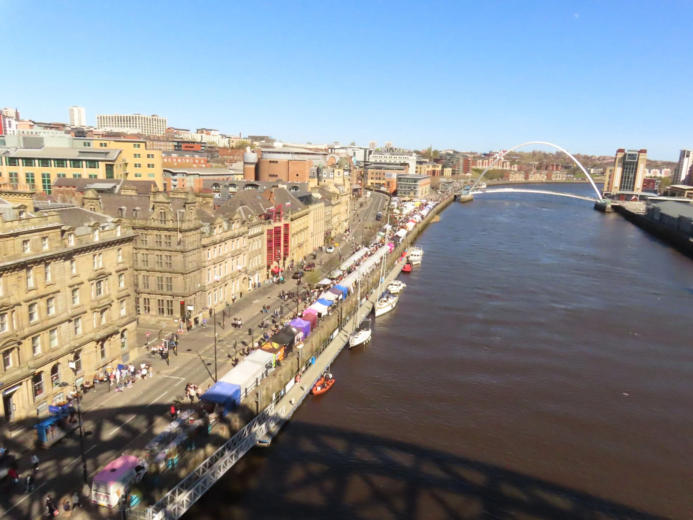Aerial view from the Tyne Bridge down the Quayside on a sunny day: a long row of colourful market stalls and crowds beside sandstone buildings, boats moored along the river bank, and the curved arch of the Gateshead Millennium Bridge across the River Tyne in the distance under a clear blue sky