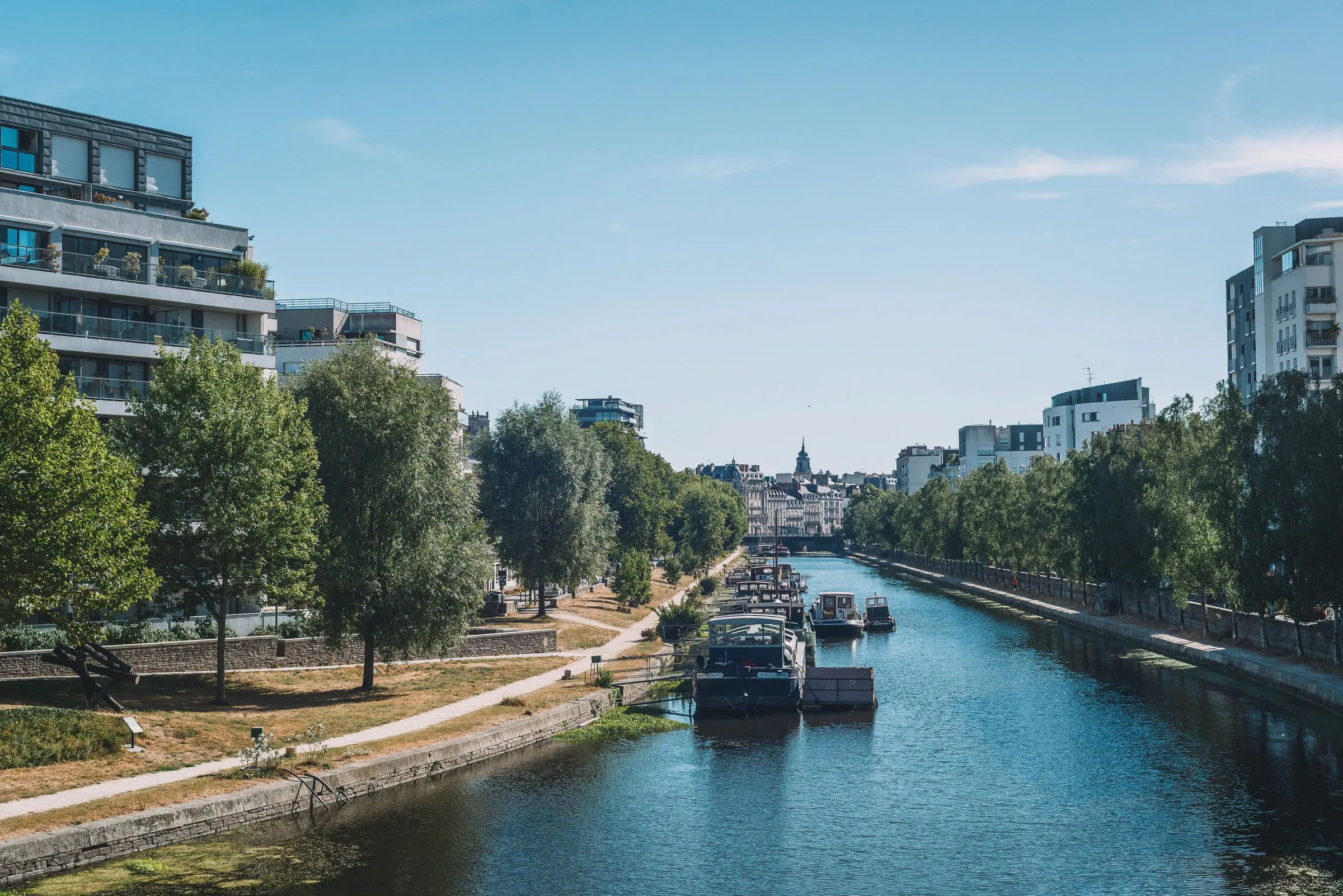 A canal in Rennes on a sunny day, with moored houseboats, a riverside footpath lined with trees and modern apartment blocks on both banks, and the old town's spires in the distance