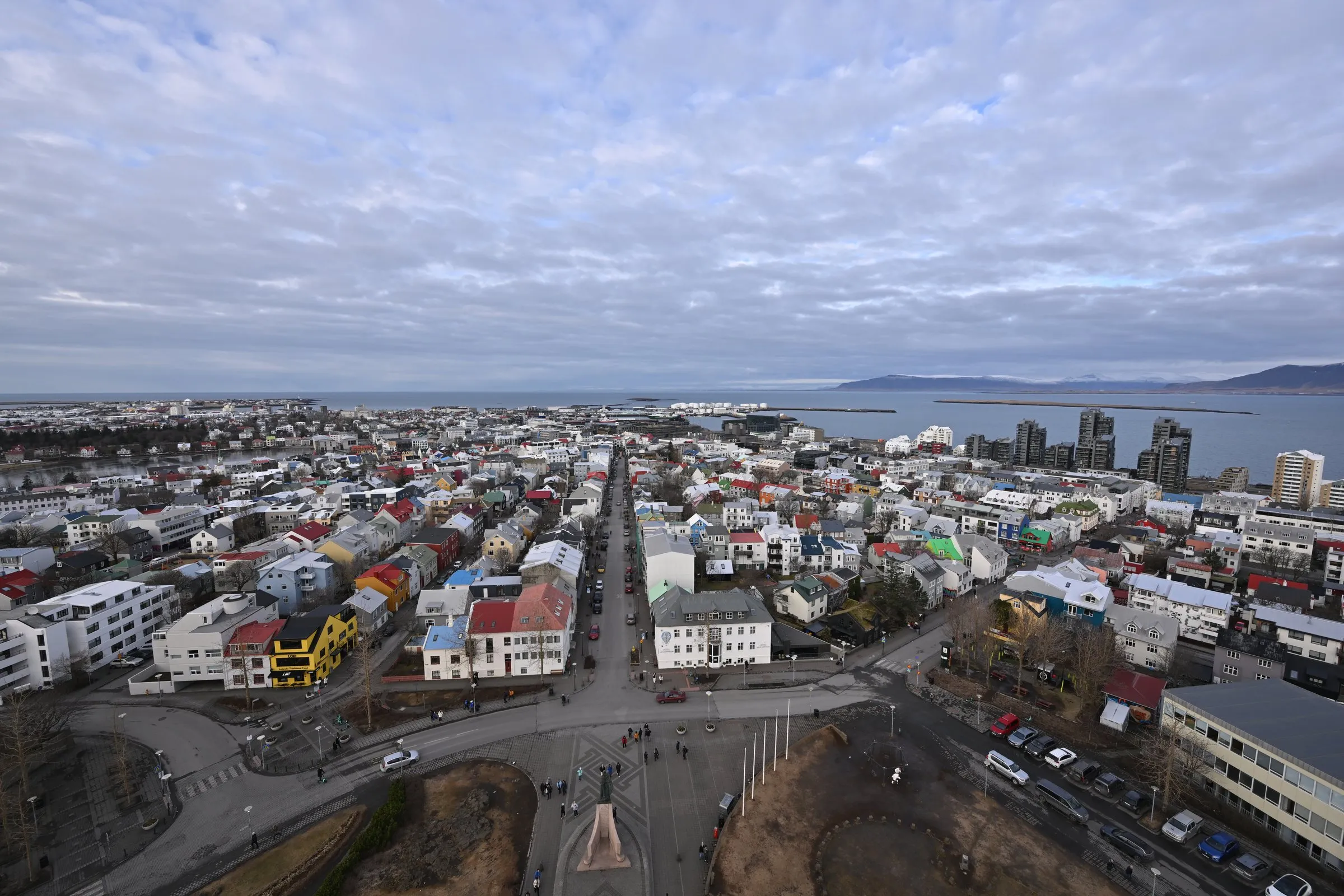 View north-west from the Hallgrímskirkja tower over Reykjavík's colourful rooftops and the Skólavörðustígur street running down to the sea, with the Leifur Eiríksson statue in the square below and mountains across the bay under a cloudy sky