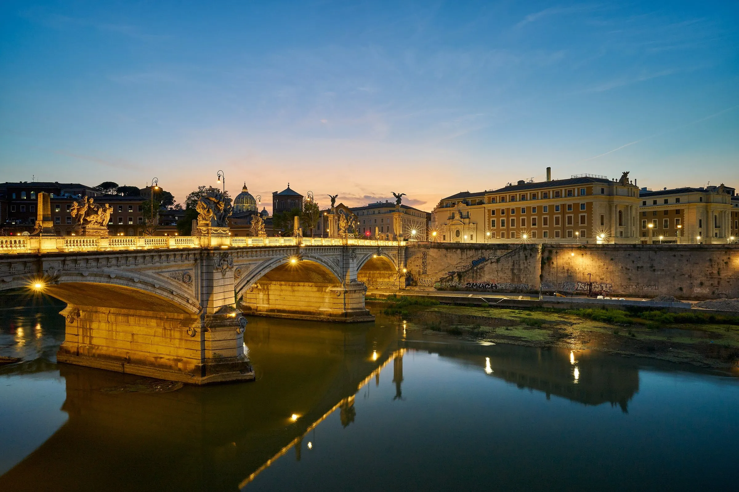 Ponte Vittorio Emanuele II over the Tiber in Rome at blue hour, its statues and arches lit up and reflected in the water, with the dome of St Peter's Basilica on the skyline behind