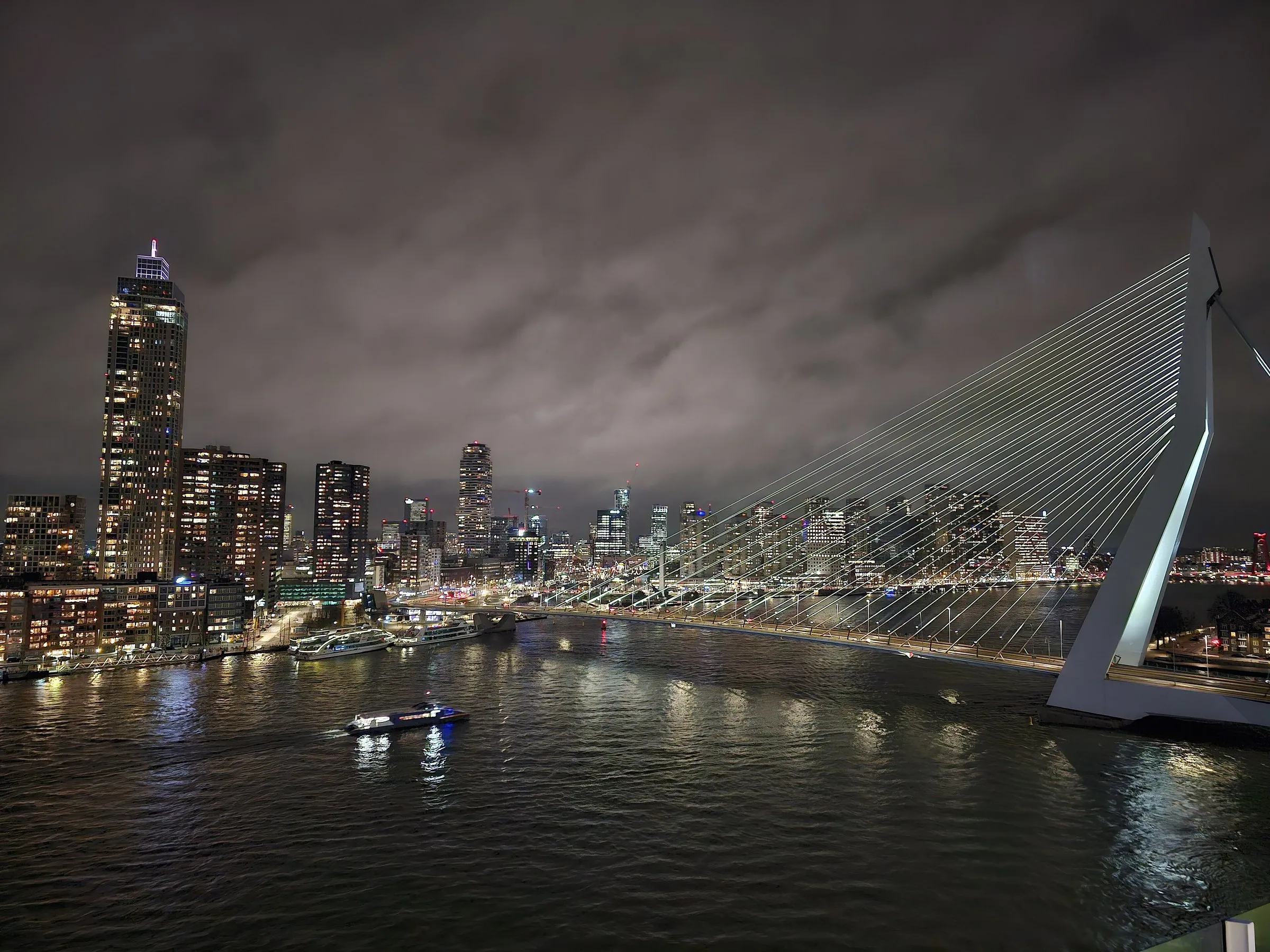 The Erasmus Bridge in Rotterdam at night under a cloudy sky, its white pylon and cables lit on the right, the lit towers of Kop van Zuid on the left and a waterbus crossing the dark Nieuwe Maas