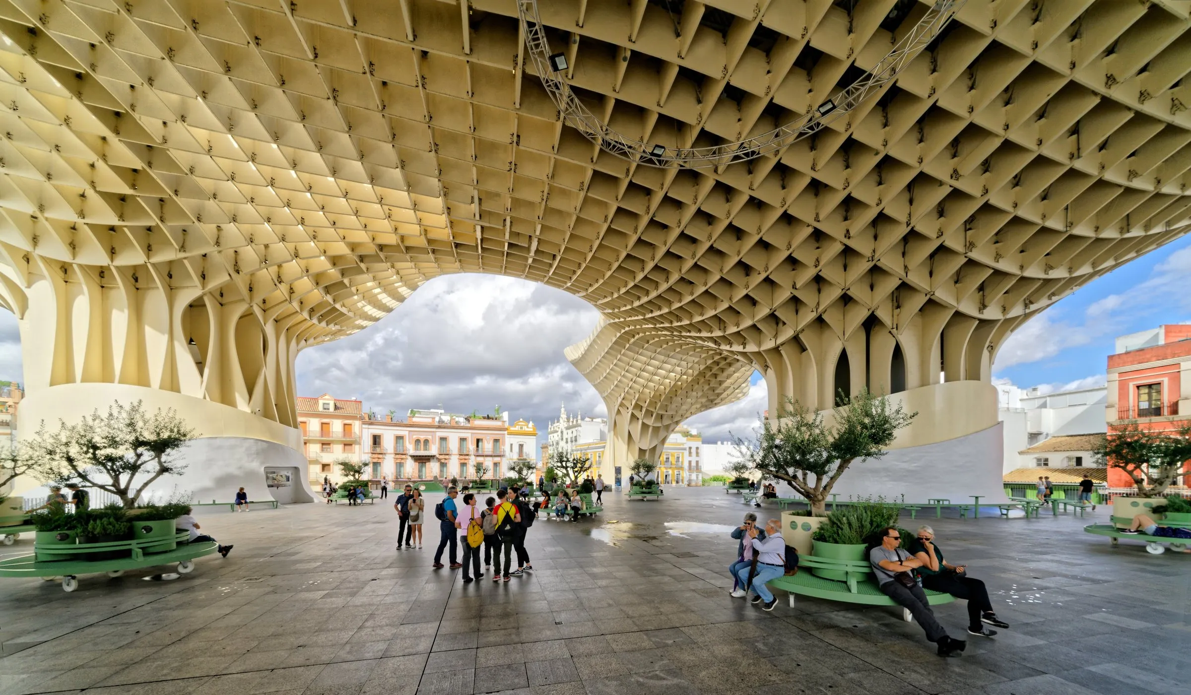 The underside of the Metropol Parasol in Seville: a pale wooden lattice canopy on curved columns over the Plaza de la Encarnación, with people walking and sitting on green benches among potted olive trees, and old buildings under a cloudy blue sky in the background