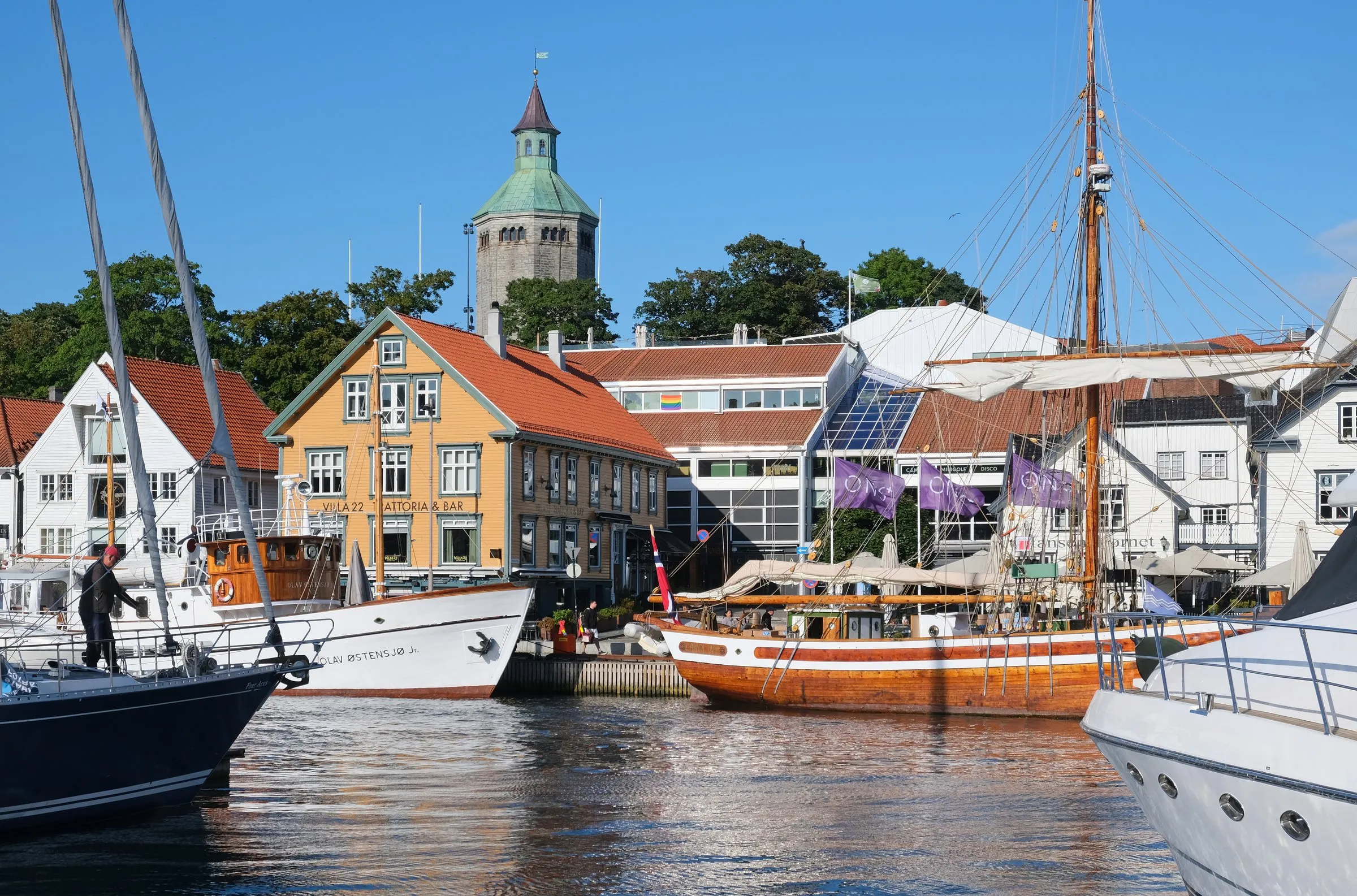 Boats and a wooden sailing ship moored at Skagenkaien in the Vågen harbour, in front of white and ochre wooden houses and purple flags, with the stone Valberg tower and its green copper roof rising behind under a clear blue sky