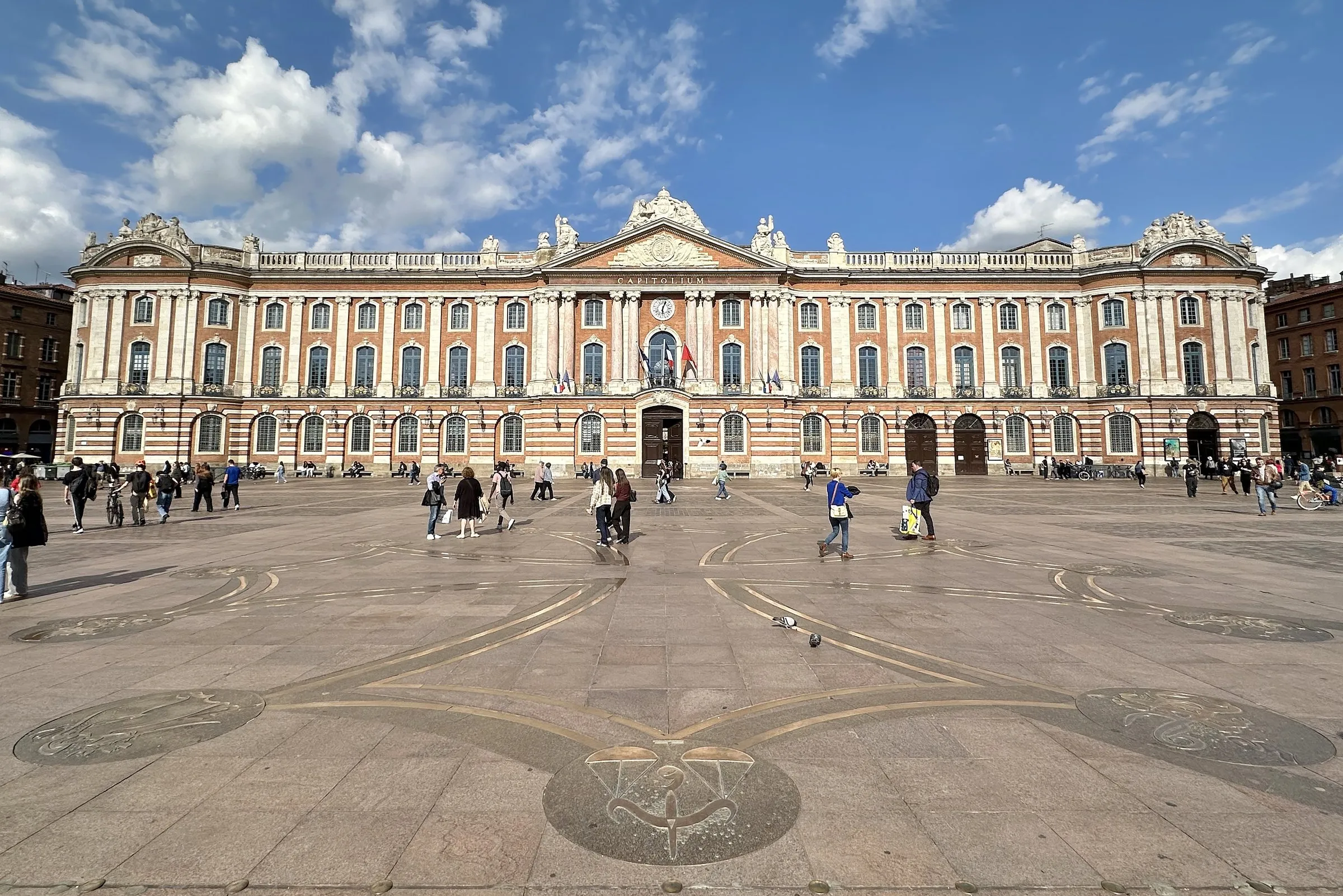 The Capitole, Toulouse's city hall, with its long pink-brick and white-stone façade under a blue sky with white clouds, seen across Place du Capitole with pedestrians and the large Occitan cross set into the paving in the foreground