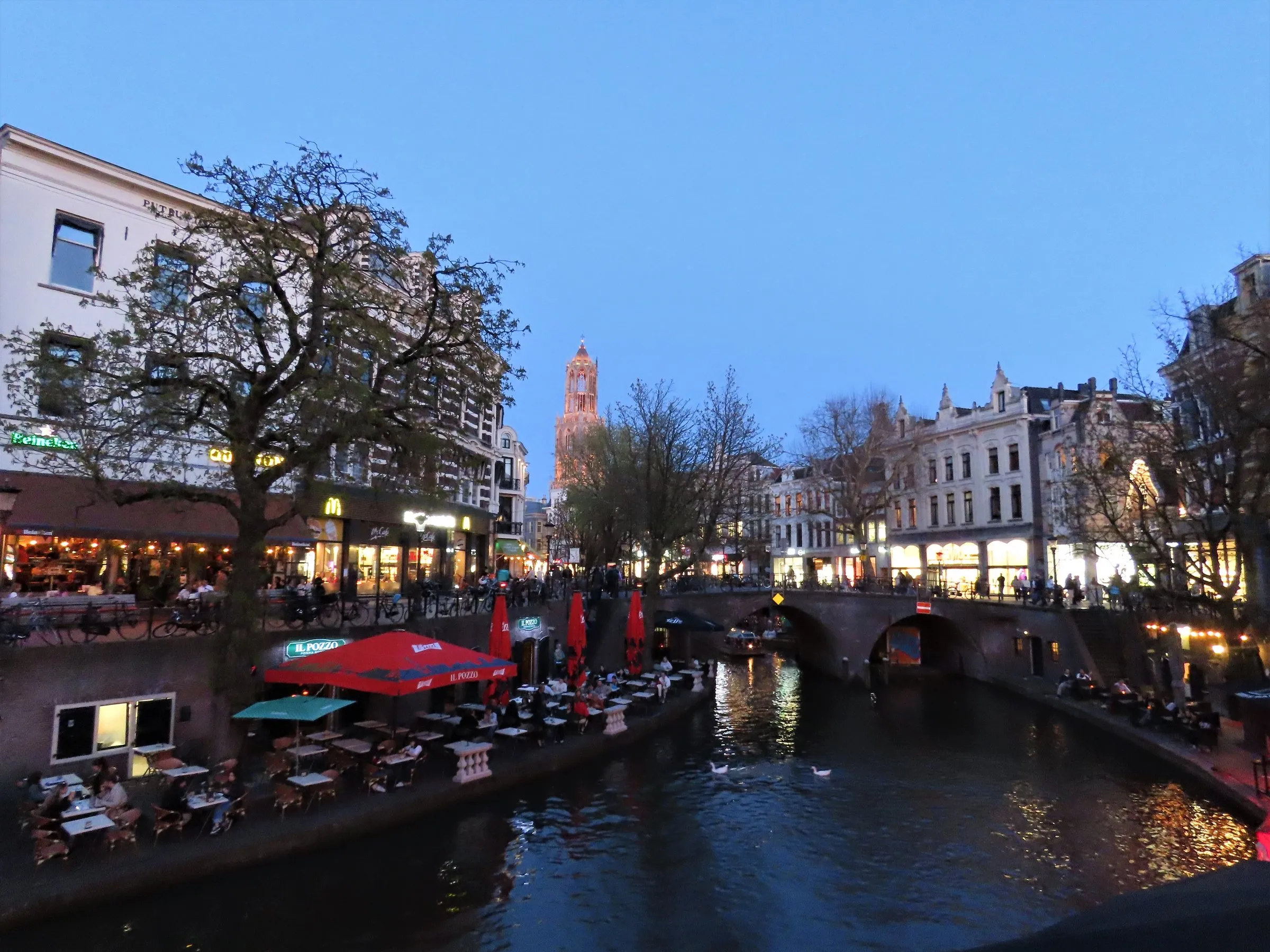 The Oudegracht canal in Utrecht at dusk, with café terraces and red parasols along the wharf, an arched brick bridge and the lit Dom tower rising behind the trees