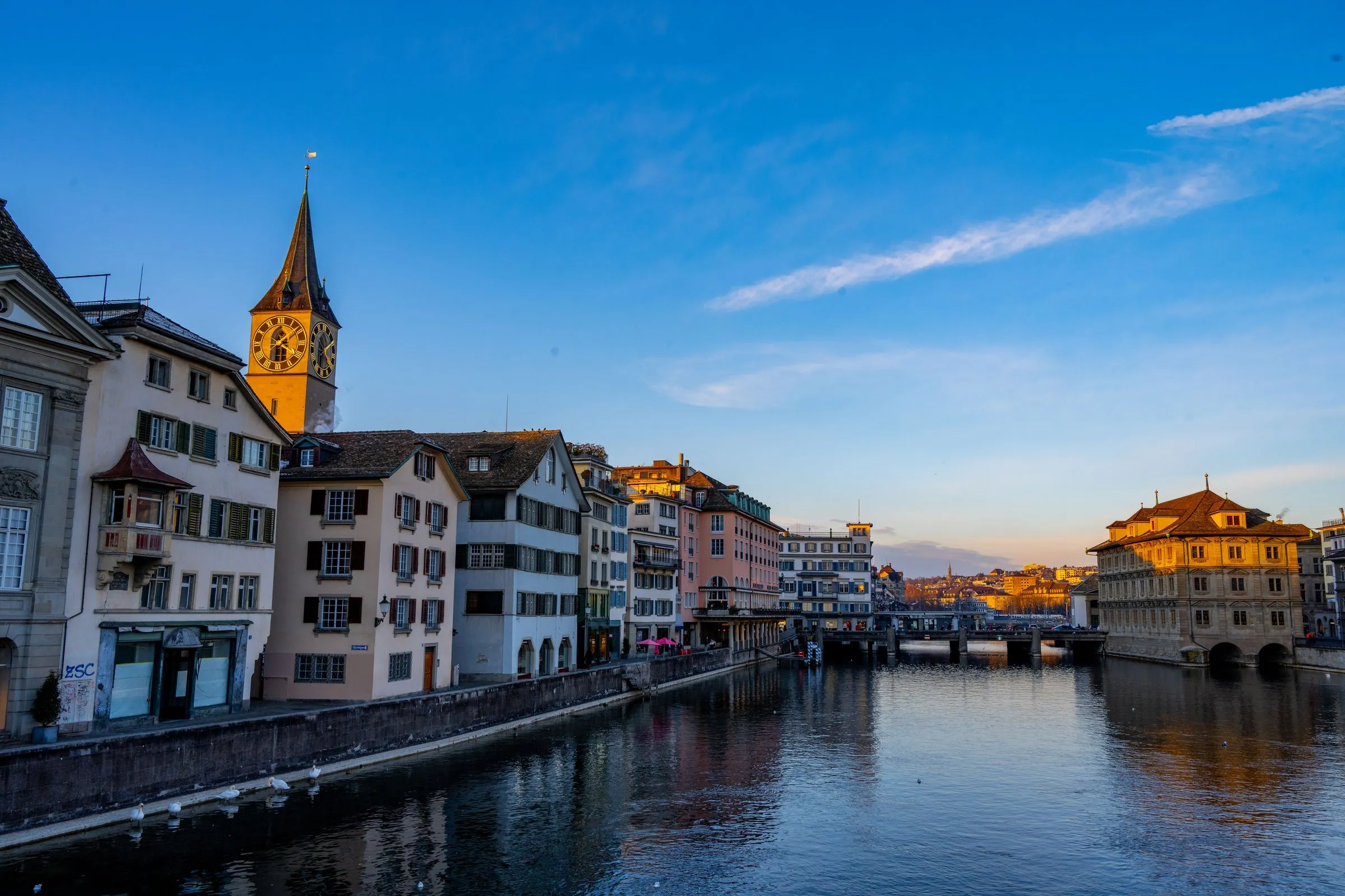 The Limmat in Zurich's old town at sunset, with the clock tower of St. Peter's Church above the riverside houses on the left, the Rathausbrücke and the Town Hall on the right