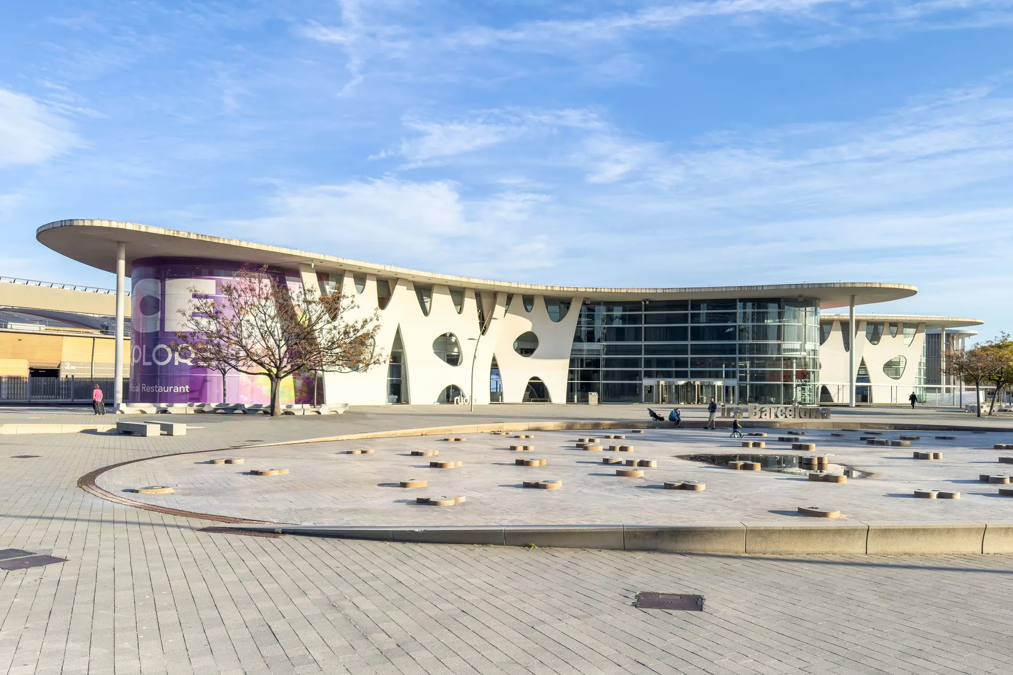 The Fira de Barcelona Gran Via venue in L'Hospitalet de Llobregat, a white building with a wide curved roof and oval openings, seen across a paved square