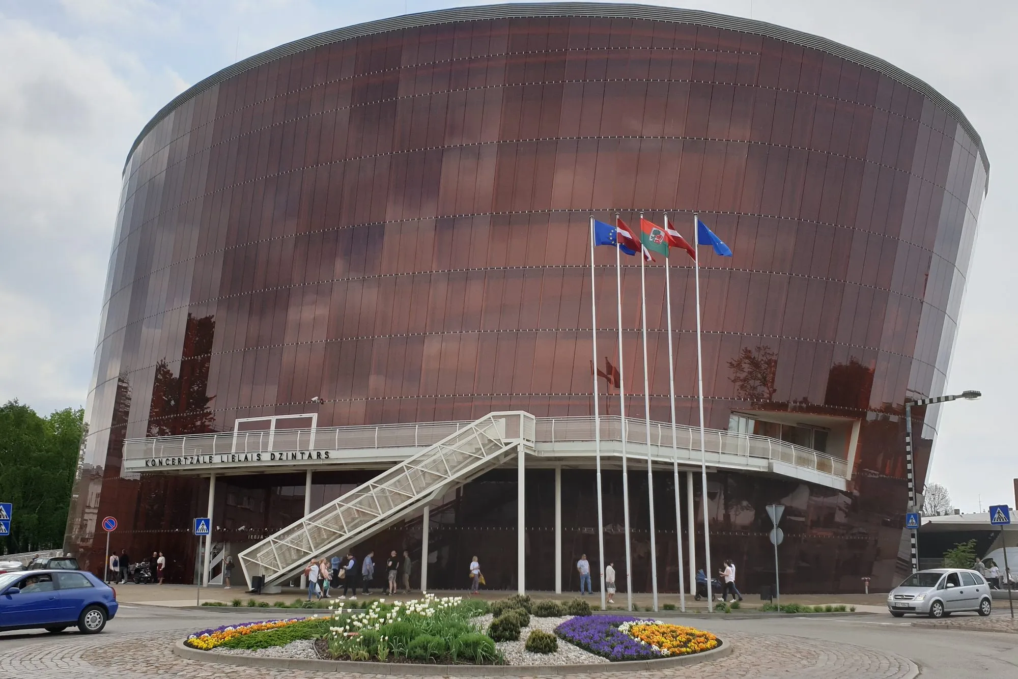The Great Amber Concert Hall in Liepāja, Latvia, a round building clad in amber-coloured glass, with flags and a flower bed in front