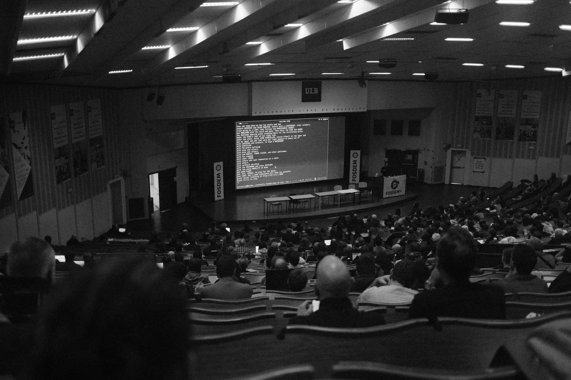 Imagine alb-negru a unui amfiteatru plin de la Université libre de Bruxelles în timpul conferinței de tehnologie FOSDEM, cu un vorbitor și un ecran mare de proiecție